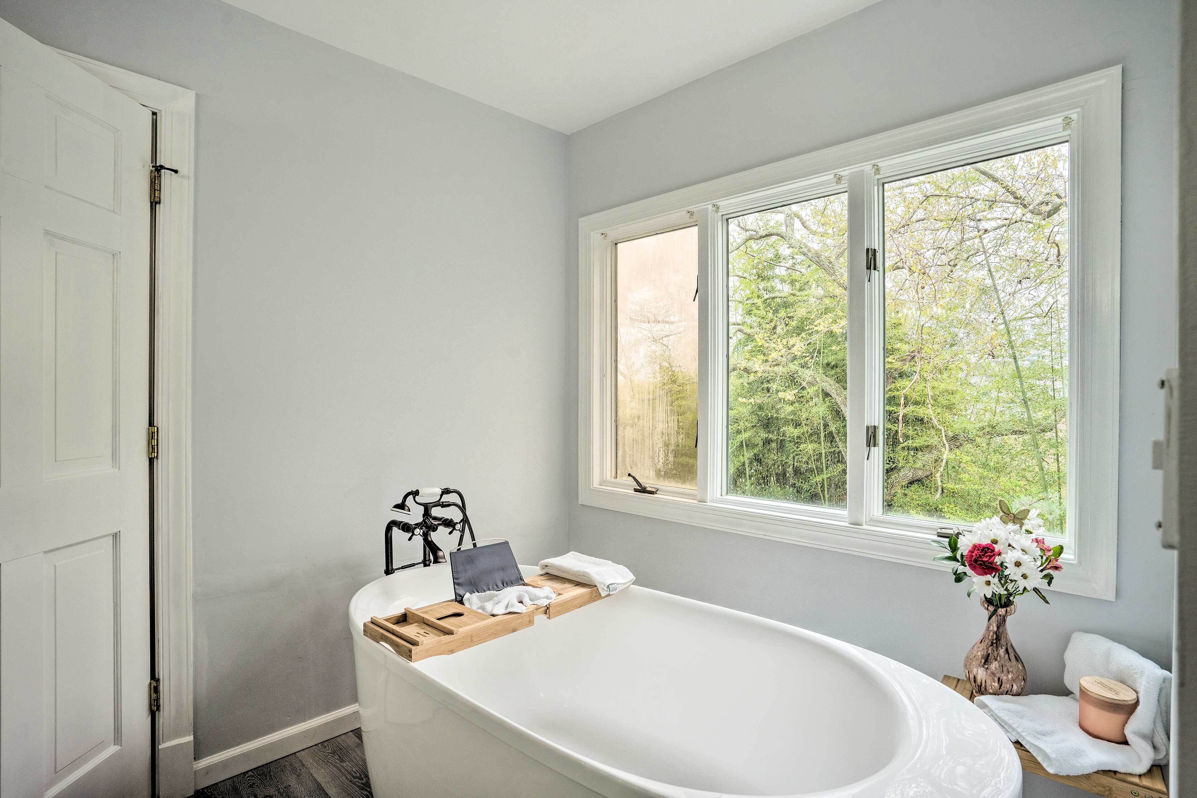 A bathroom with a freestanding bathtub, a window with a view of greenery, and a white door.