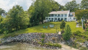 A large, two-story house with a well-maintained lawn and a stone retaining wall near a body of water. - Hot Tub + Private Beach: Waterfront Urbanna Escape (Urbanna)