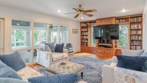 A spacious living room with a large bookshelf, a television, and a ceiling fan. - Hot Tub + Private Beach: Waterfront Urbanna Escape (Urbanna)