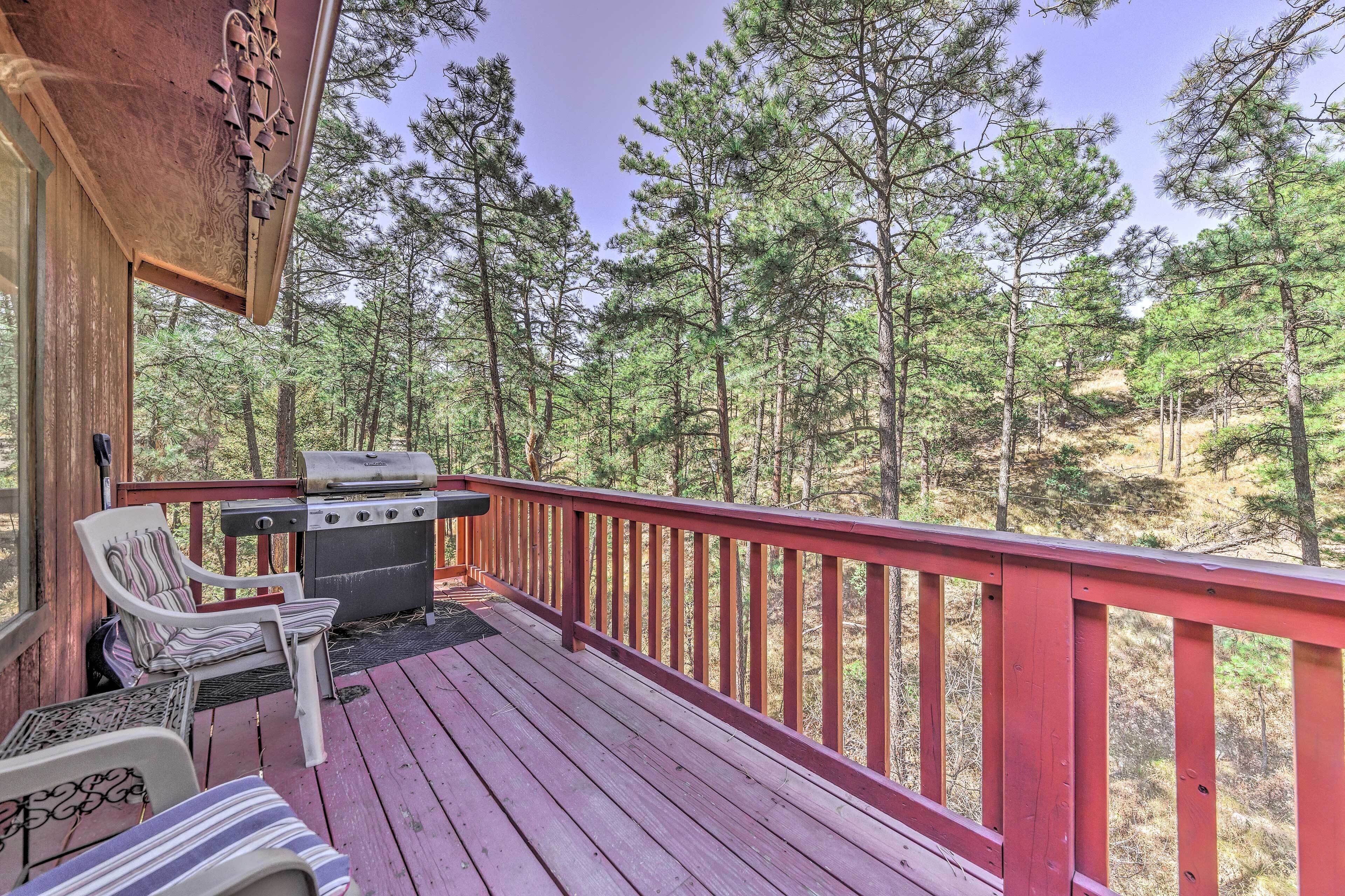 A wooden deck with a grill and two chairs, overlooking a forest.