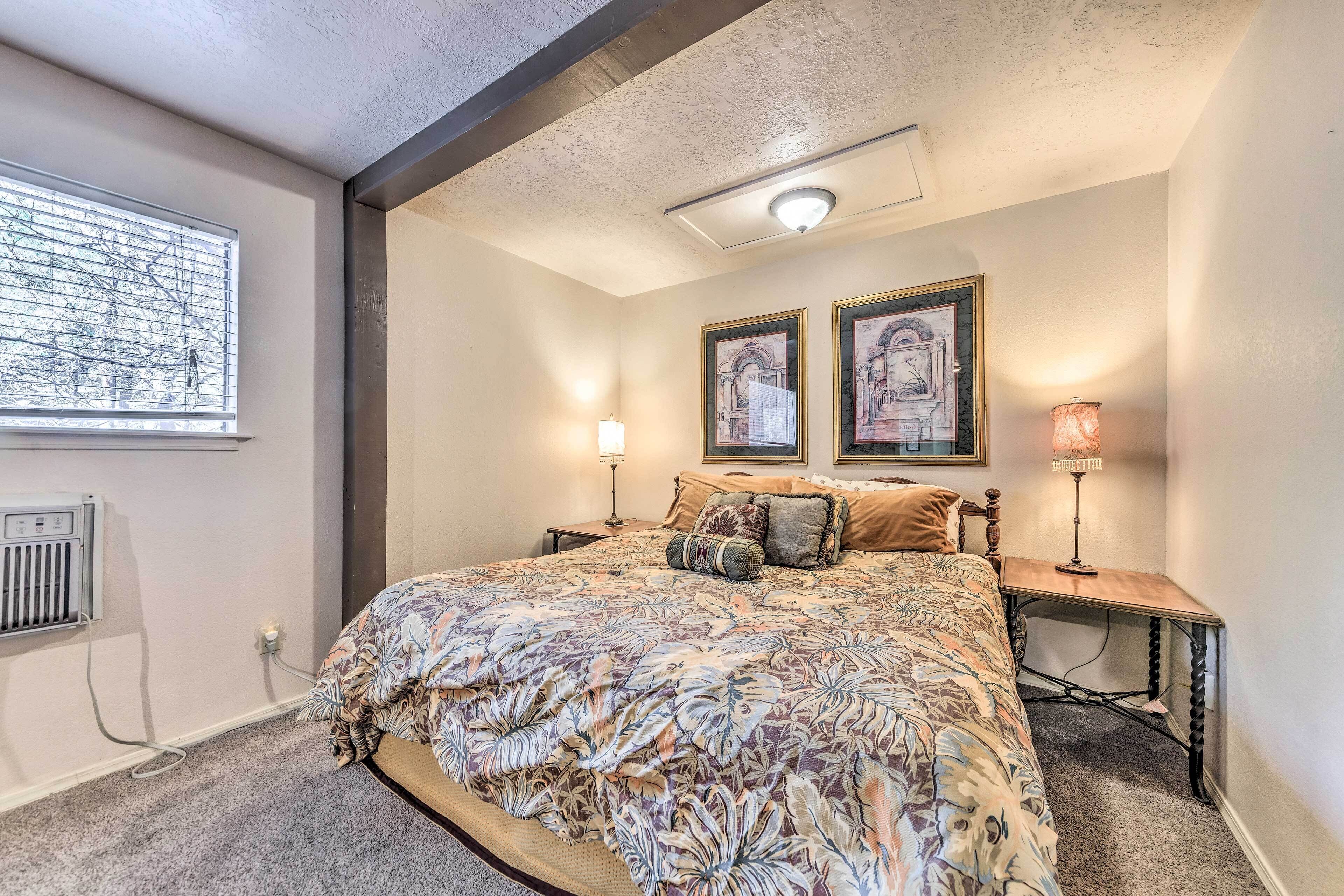 A bedroom with a large bed, two bedside tables, a window with blinds, and two framed pictures on the wall.