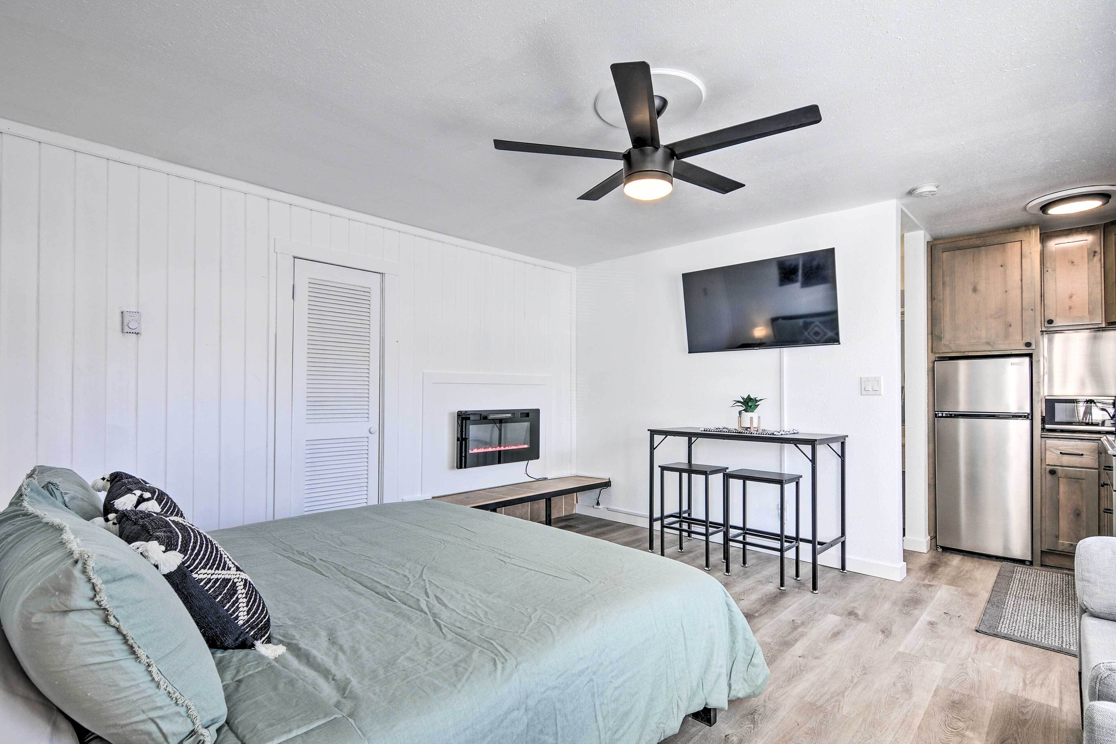 Un dormitorio moderno con cama, ventilador de techo, televisor montado en la pared y una pequeña cocina equipada con refrigerador y estufa.