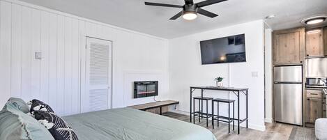 Un dormitorio moderno con cama, ventilador de techo, televisor montado en la pared y una pequeña cocina equipada con refrigerador y estufa.
