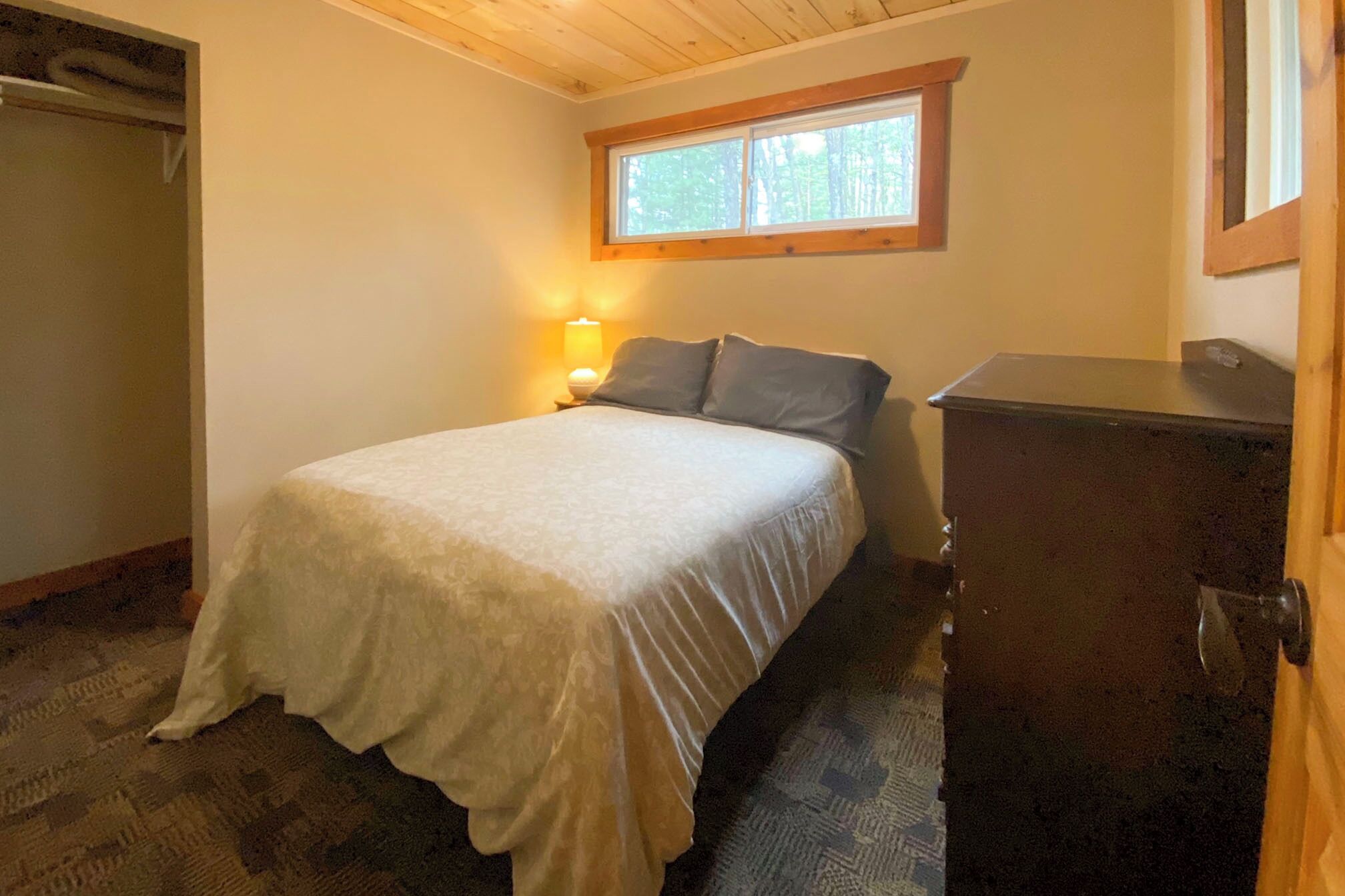 A bedroom with a bed, a nightstand with a lamp, a window, and a dresser.