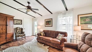 A room with a brown leather sofa, a wooden cabinet, a floral armchair, a white cowhide rug, and a ceiling fan. - On Scenic 60-acre Farm: Carmine Cottage w/ Porch (Carmine)