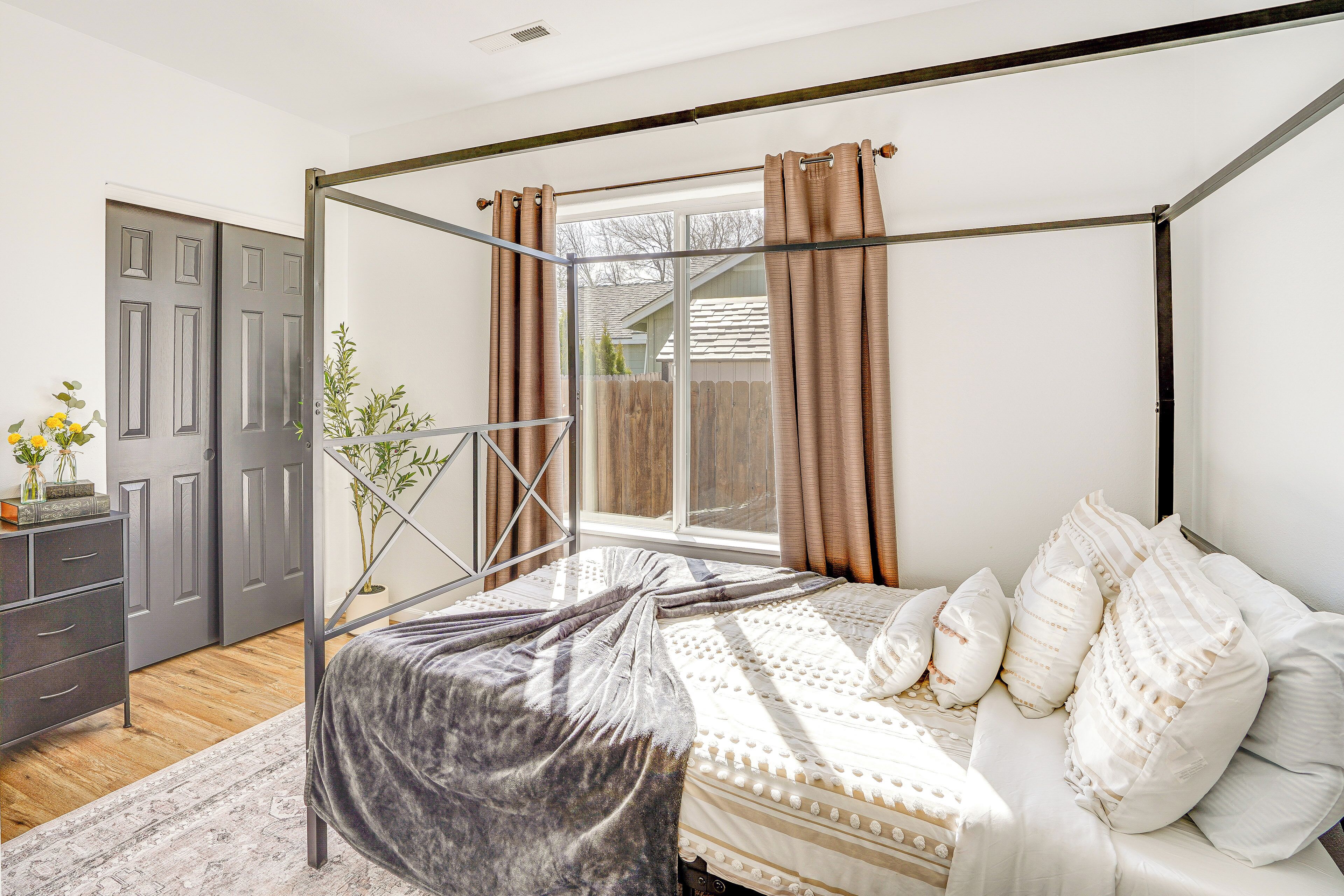 A four-poster bed with a grey bedspread, white pillows, and a window with curtains.