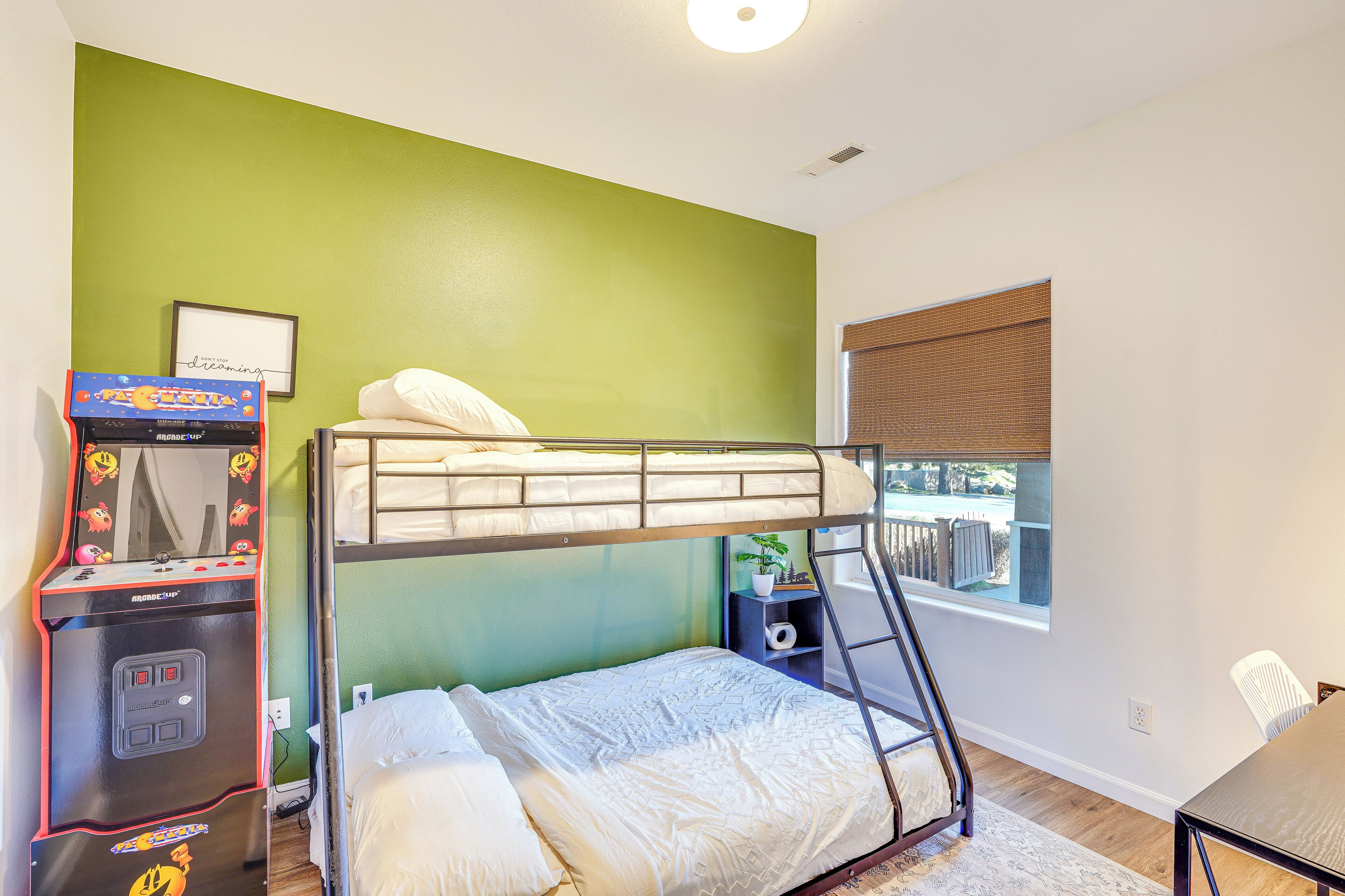 A bunk bed room with a green wall, a window with blinds, a small desk, and a gaming console.