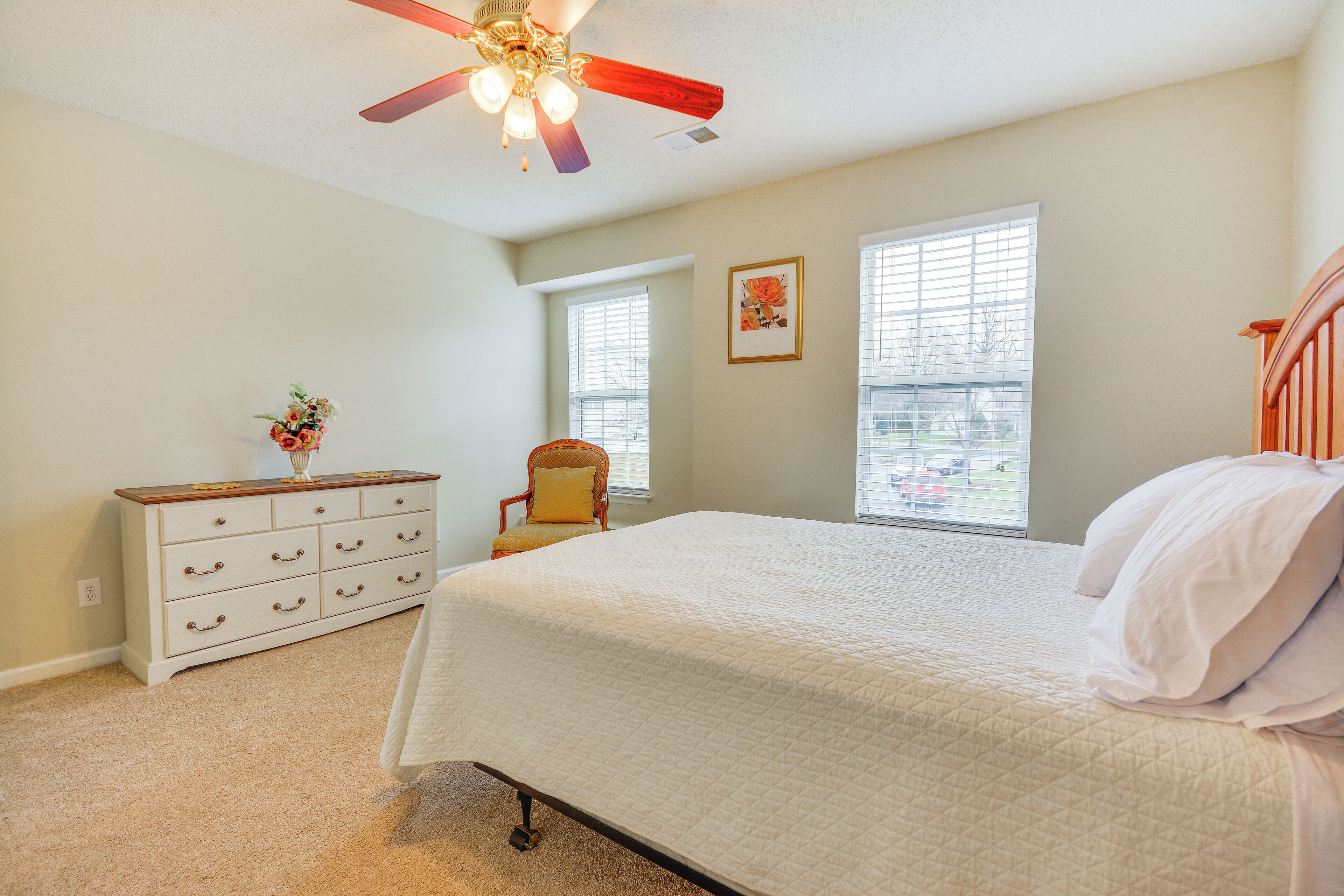 A bedroom with a bed, dresser, chair, windows, and ceiling fan.