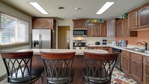 A kitchen with wooden cabinets, a stainless steel refrigerator, a microwave, and a dishwasher. There are three bar stools and a window with blinds. - Chic Silver City Getaway w/ Patio + Gas Grill (Silver City)