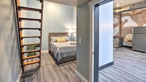 Une chambre avec un lit, une échelle en bois et une vue sur un espace de vie doté d’un mur en briques et d’un canapé.