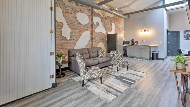 Un salon moderne avec un canapé, deux fauteuils à motifs, une table basse et un tapis. On peut voir un mur en briques dont l’isolant et les conduits sont apparents.