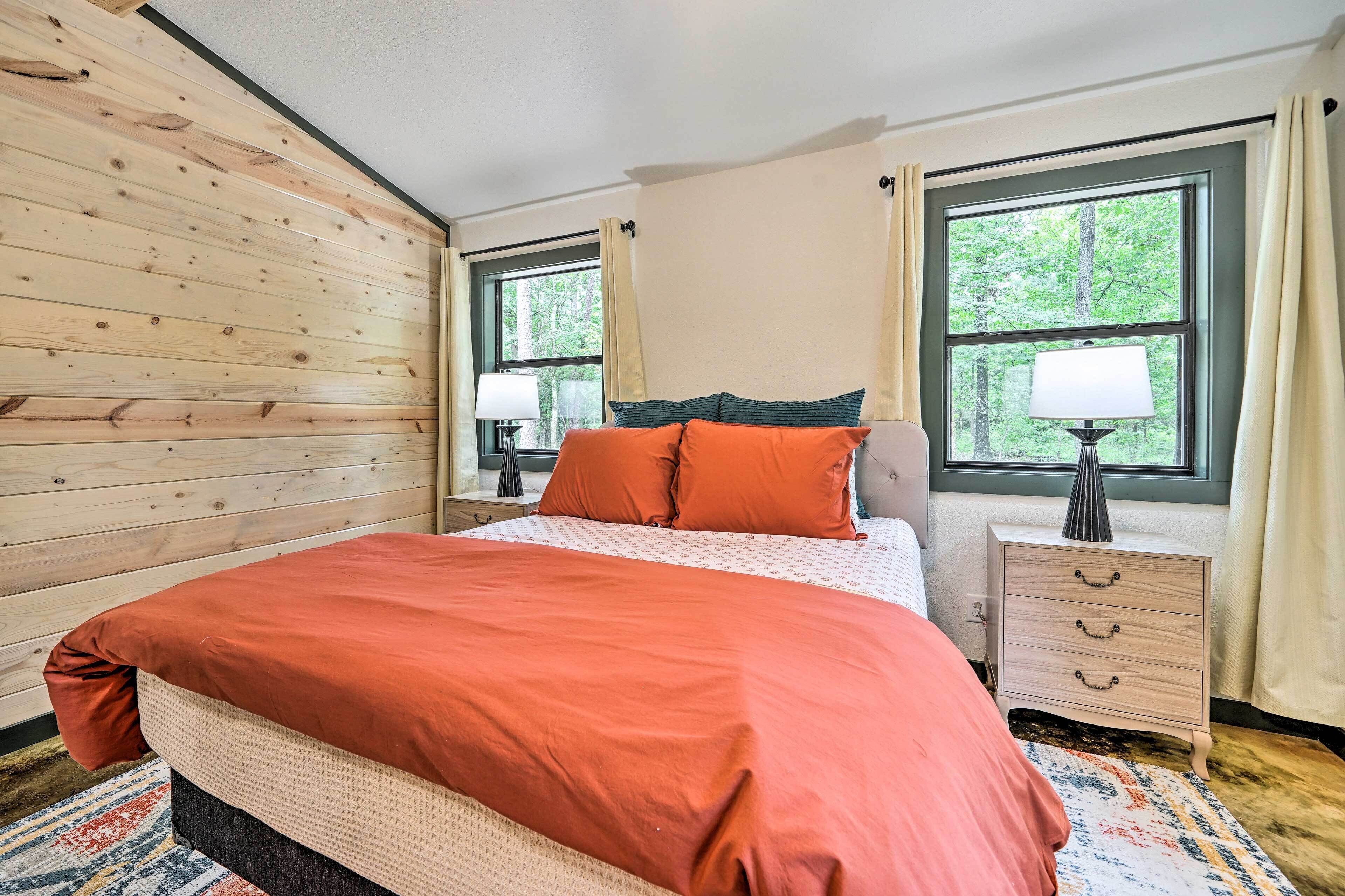 A bedroom with a wooden slat wall, a bed with orange bedding, two bedside tables with lamps, and windows with curtains.