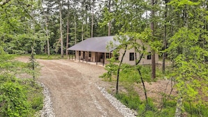 A rustic cabin with a gravel driveway leading to a covered porch, surrounded by dense forest. - Quiet & Peaceful Hochatown Cabin w/ Hot Tub! (Broken Bow)