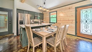A modern kitchen with a central island, wooden cabinetry, and a dining area with a table set for four. - Quiet & Peaceful Hochatown Cabin w/ Hot Tub! (Broken Bow)