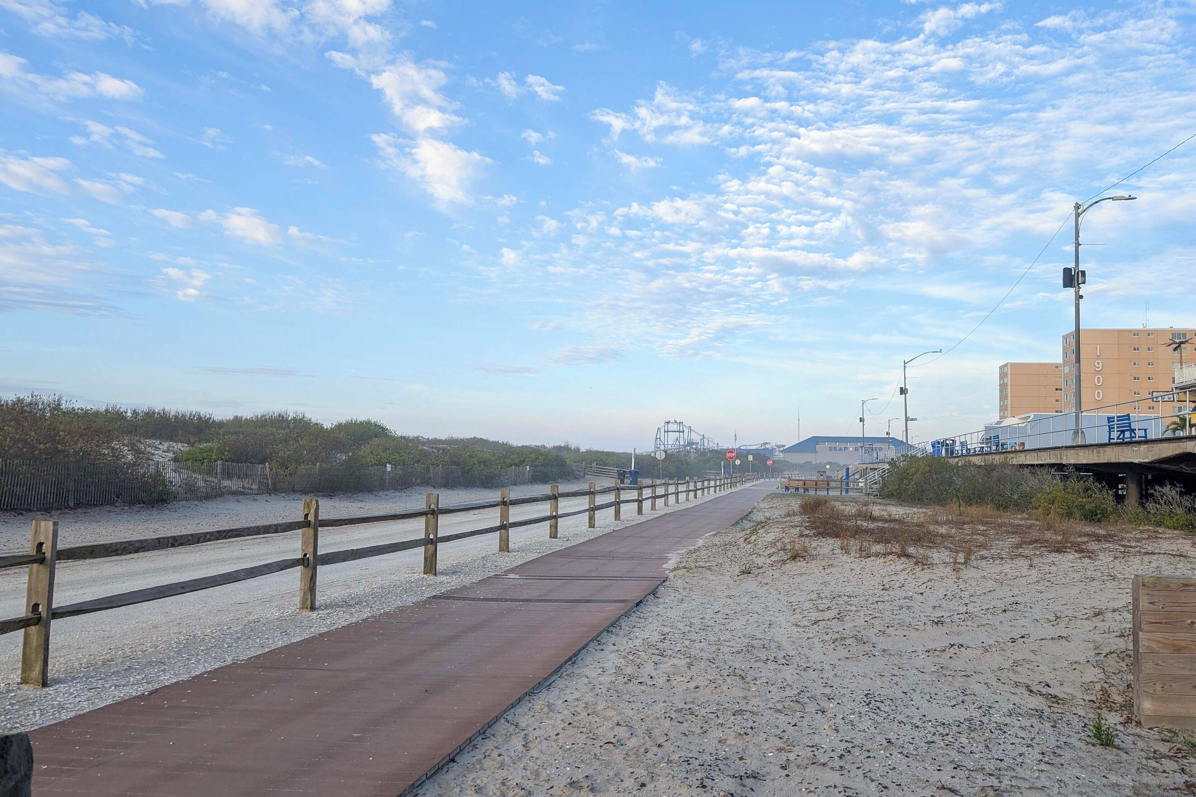 Un paseo marítimo con valla de madera, sendero arenoso y un edificio al fondo.