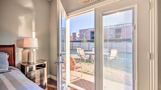 A bedroom with a bed, nightstand, lamp, and a sliding glass door leading to an outdoor patio with chairs and a table. - Cozy Duplex, Walk to Bentonville City Square! (Bentonville)