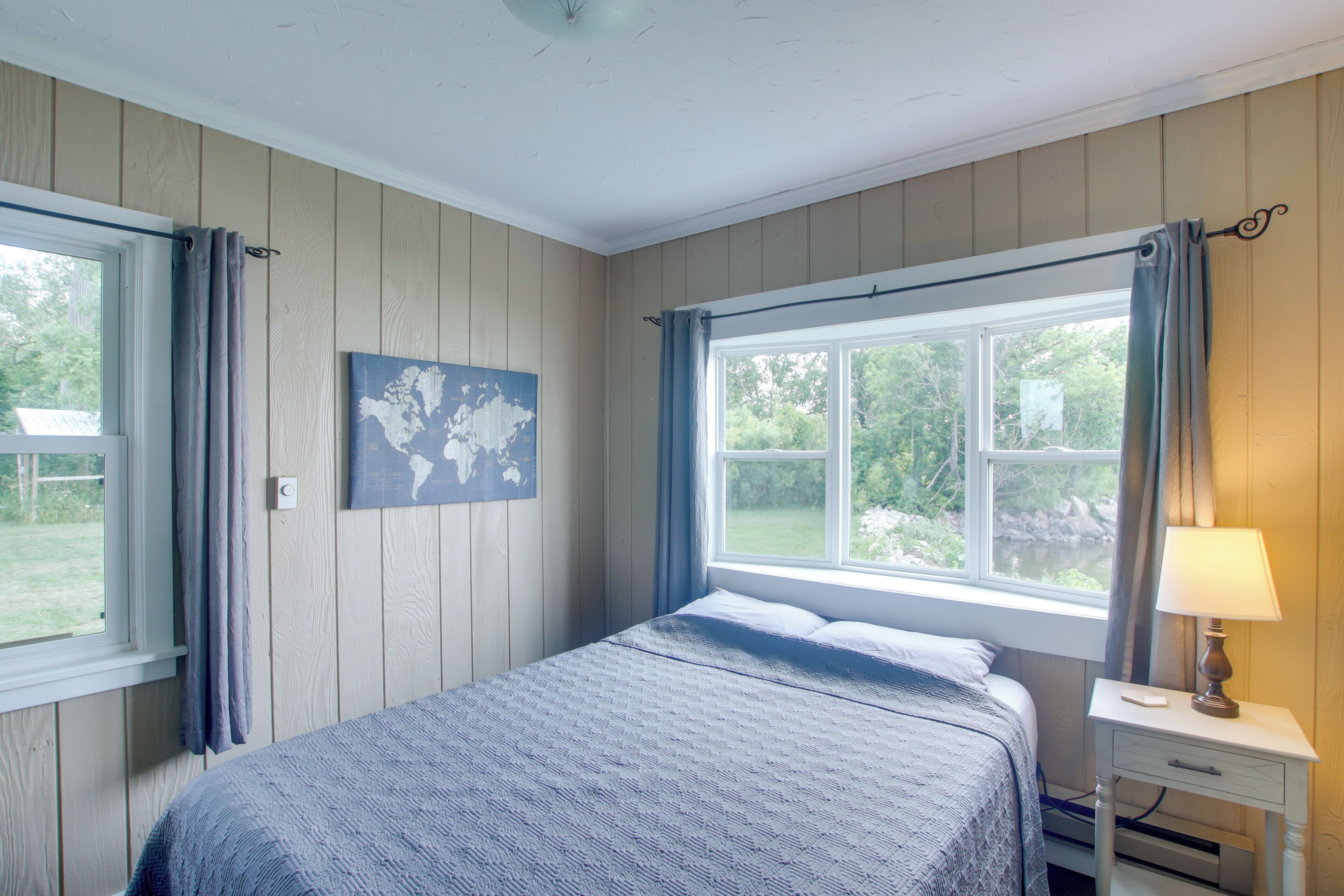 A bedroom with a bed, a nightstand, a lamp, a window with curtains, and a world map on the wall.