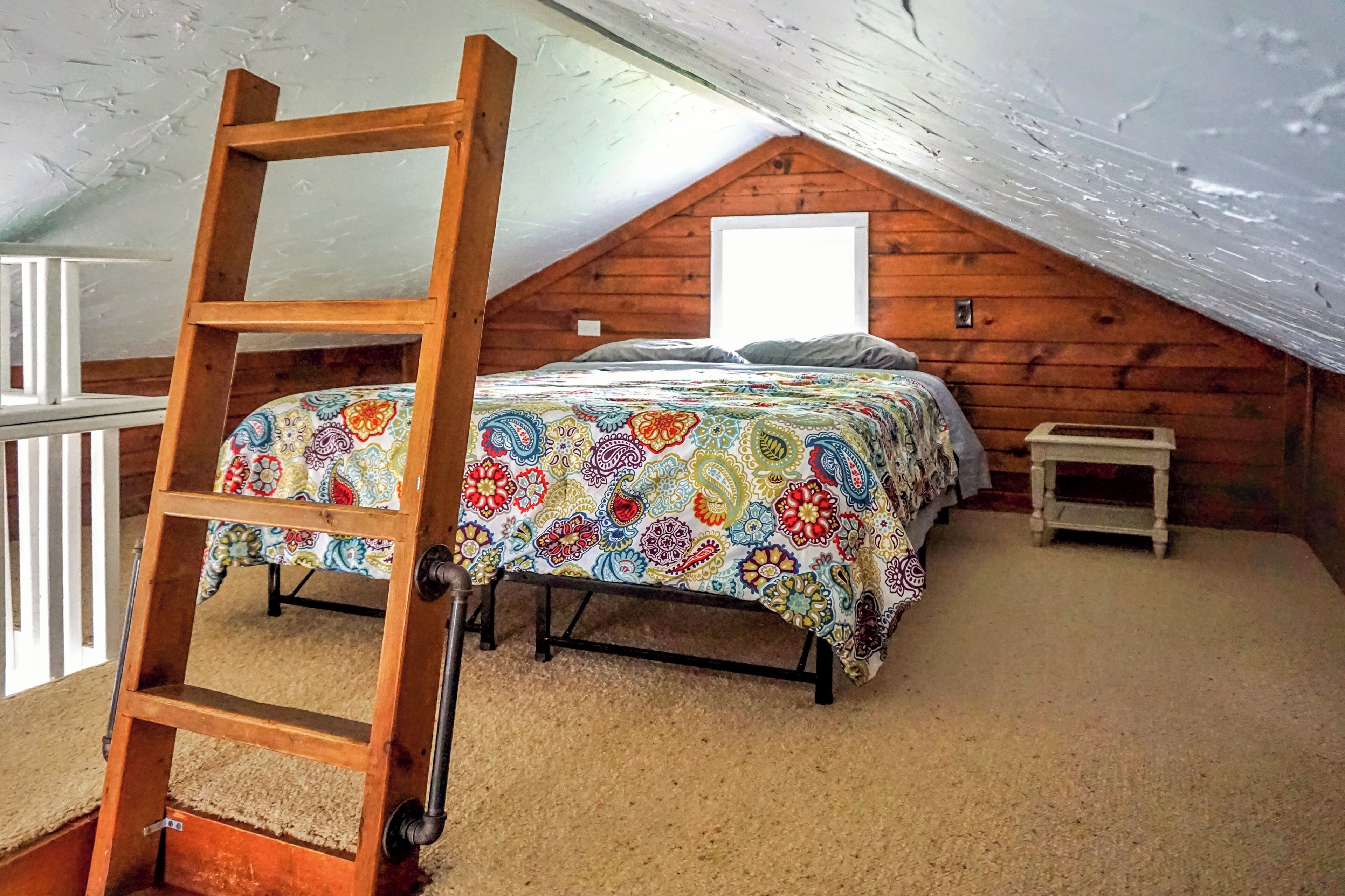 Quarto com teto estilo loft, escada de madeira, cama com colcha estampada e mesa de cabeceira pequena.