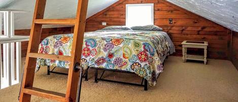 Quarto com teto estilo loft, escada de madeira, cama com colcha estampada e mesa de cabeceira pequena.