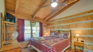 A wooden cabin bedroom with a bed, a nightstand, a fan, a TV, and a window with curtains. - Covered Deck & Hot Tub: Cozy Gatlinburg Cabin (Gatlinburg)