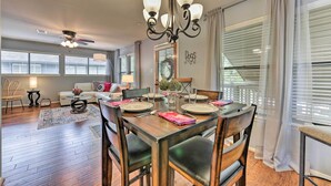 A dining area with a wooden table set for four, surrounded by chairs. A chandelier hangs above, and the room has large windows with blinds.