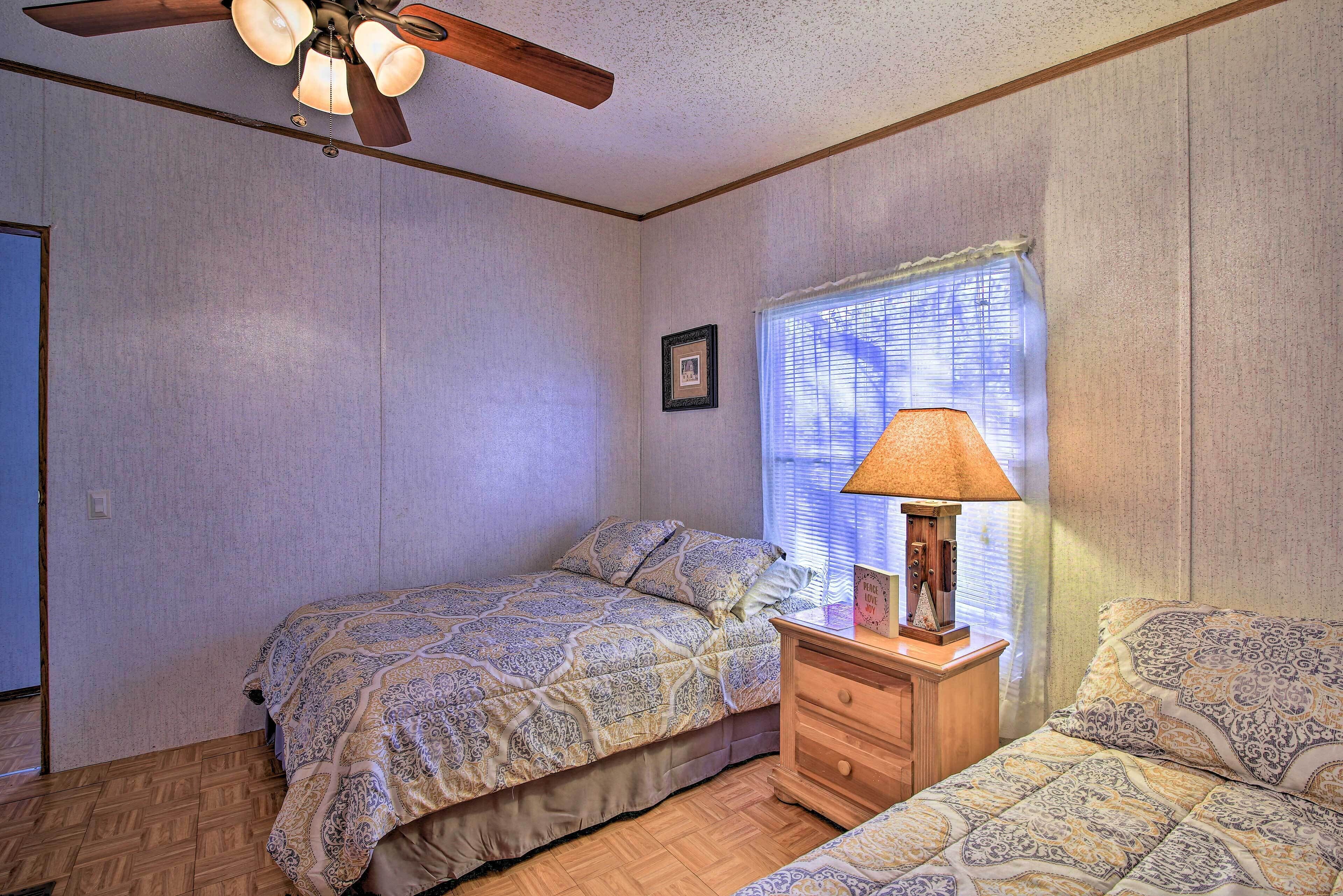 A bedroom with two beds, a ceiling fan, a window with blinds, and a bedside table with a lamp.
