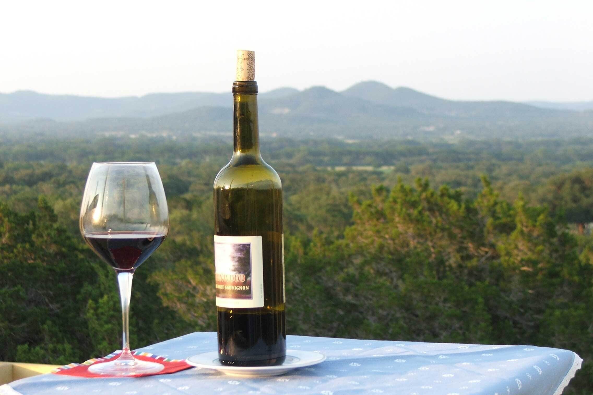 A bottle of wine and a glass on a table with a scenic view of mountains and greenery in the background.
