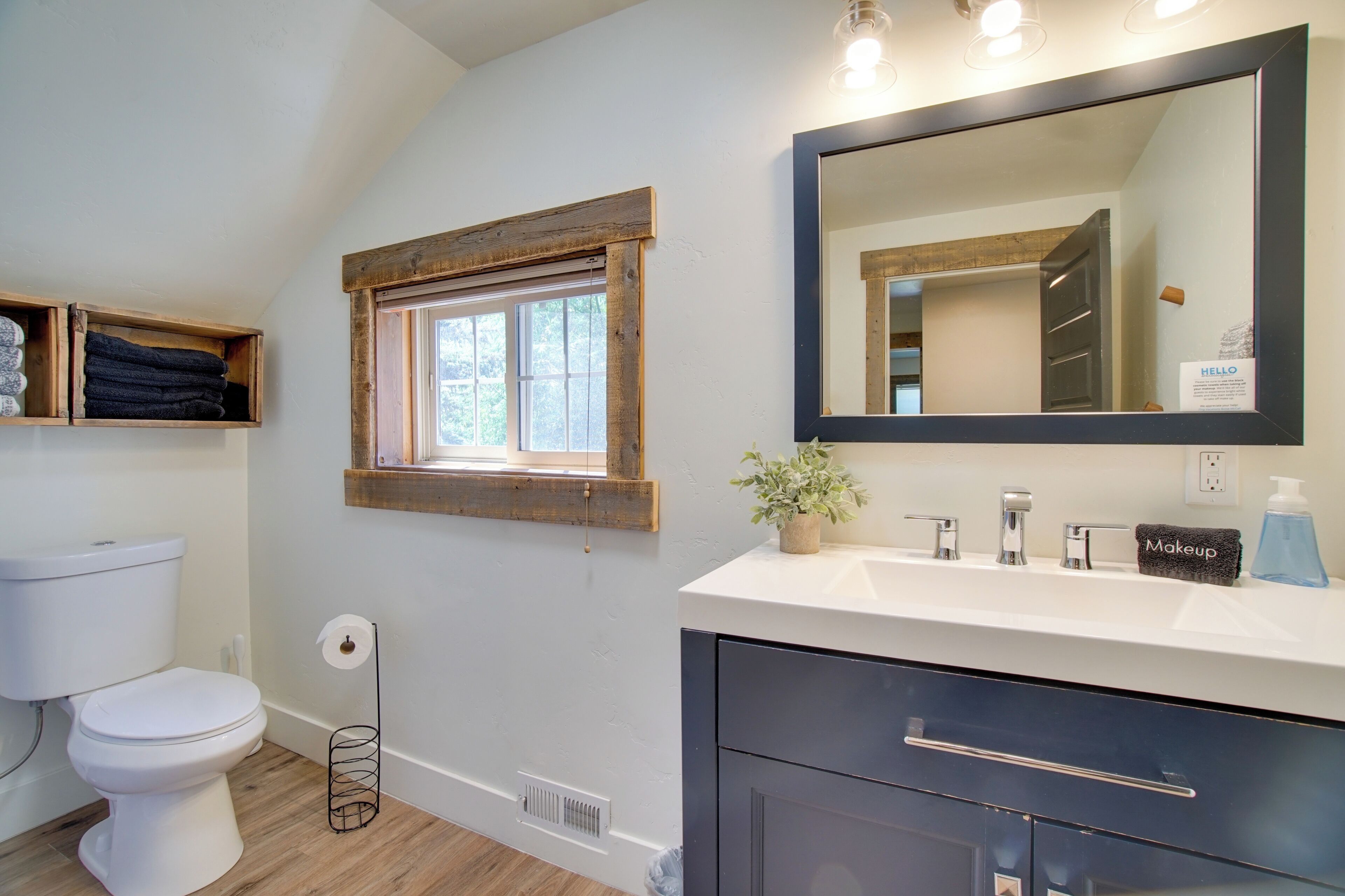A bathroom with a white toilet, wooden shelves, a window, a sink with a mirror, and a towel rack.