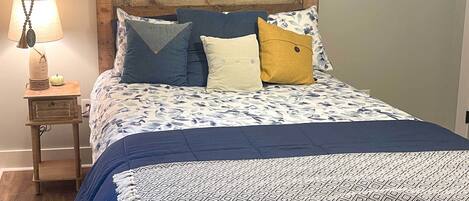 A neatly made bed with a patterned bedspread and multiple pillows. There is a small wooden nightstand with a lamp beside the bed.