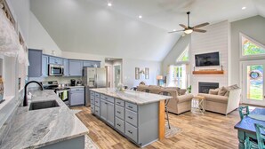 A spacious kitchen with a large island, grey cabinets, and a ceiling fan. There is a living area with a sofa and a fireplace. - 2 Mi to Golf Course + Fire Pit: Cozy Counce Cabin! (Counce)