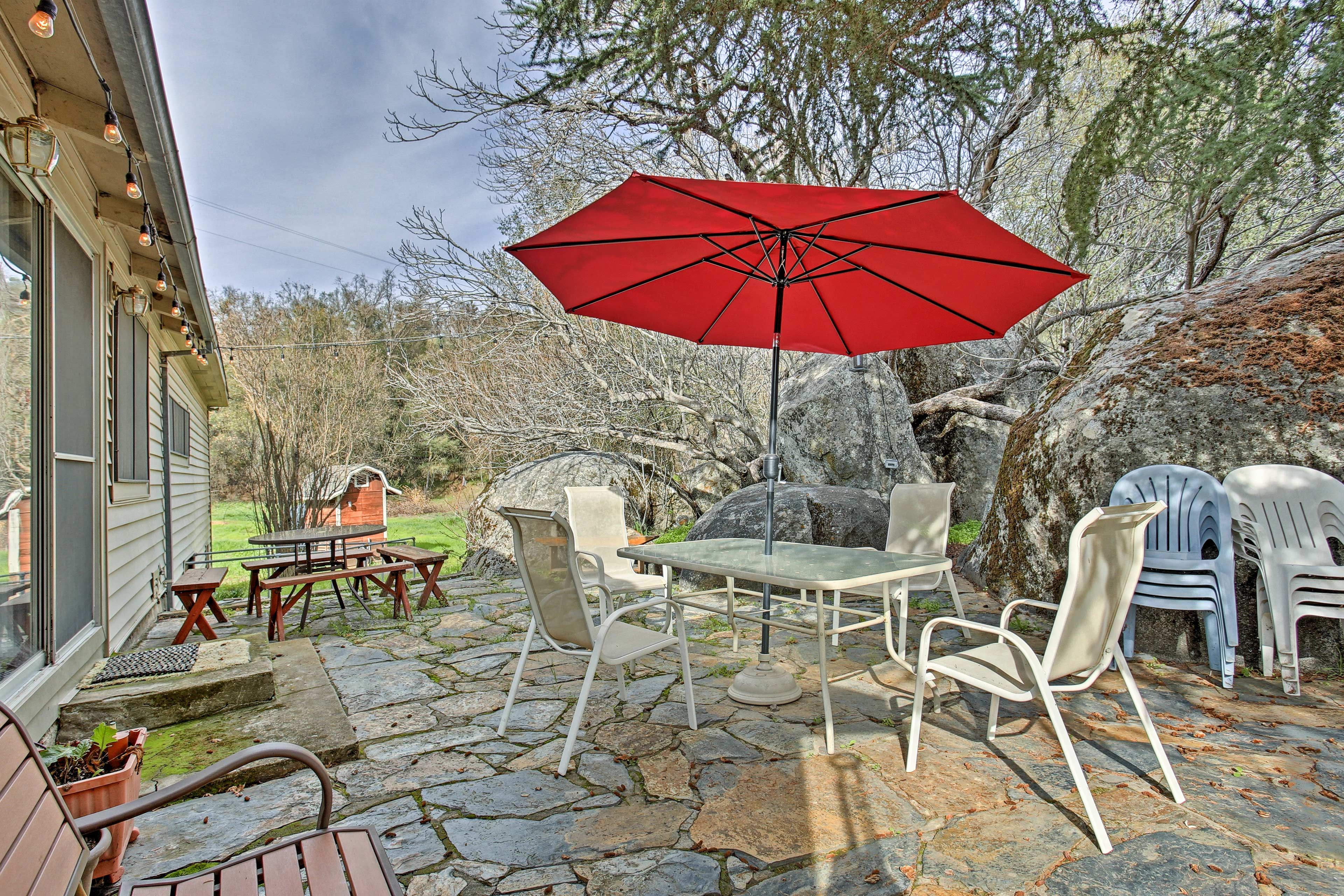 Une terrasse avec un sol en pierre, une table et des chaises, un parasol rouge et un petit abri de jardin.