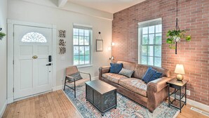 A cozy living room with a brick wall, a white door, a brown leather sofa, a patterned rug, and a small side table with a lamp.
