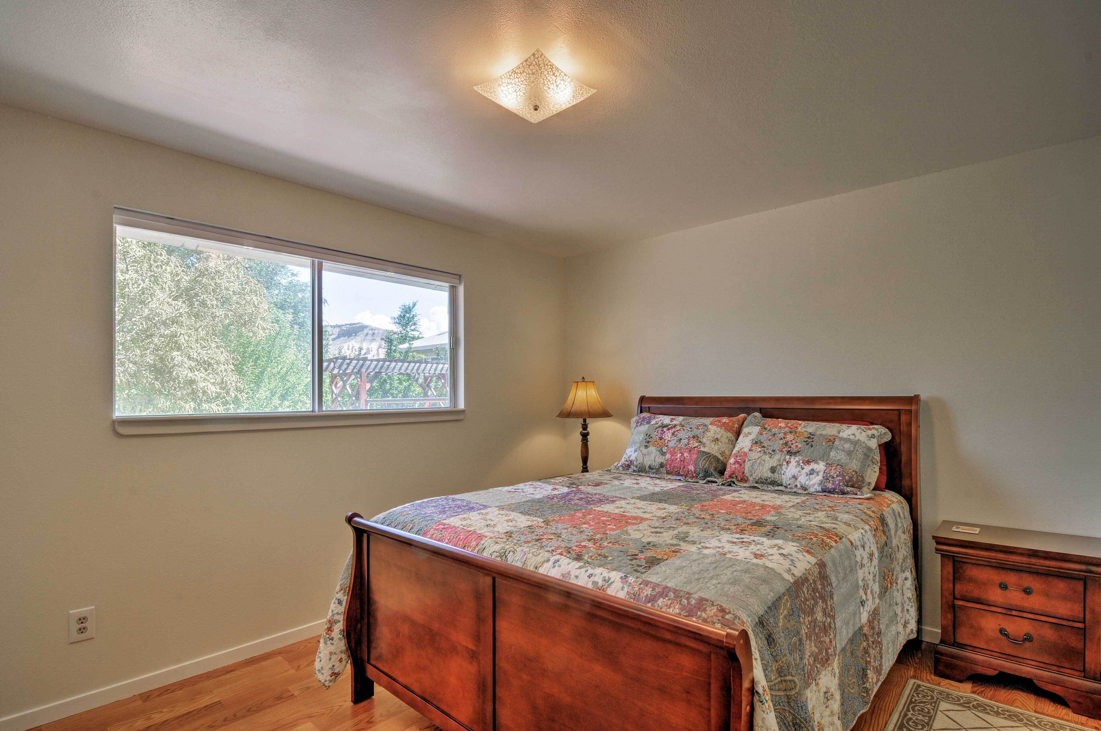 A bedroom with a wooden bed, a nightstand, a lamp, and a window with a view of trees and buildings.