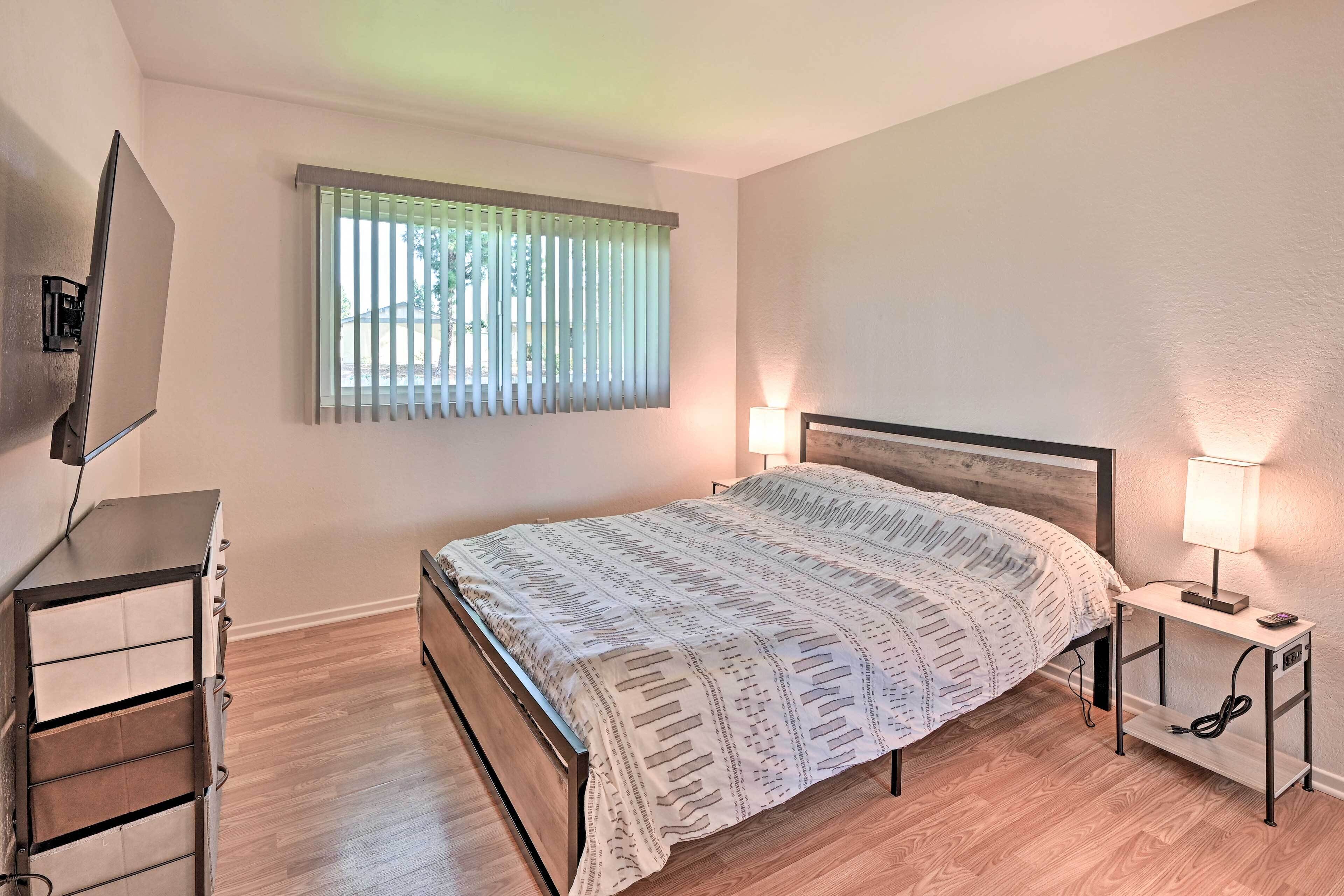 A bedroom with a bed, a bedside table with a lamp, a TV mounted on the wall, and a window with blinds.