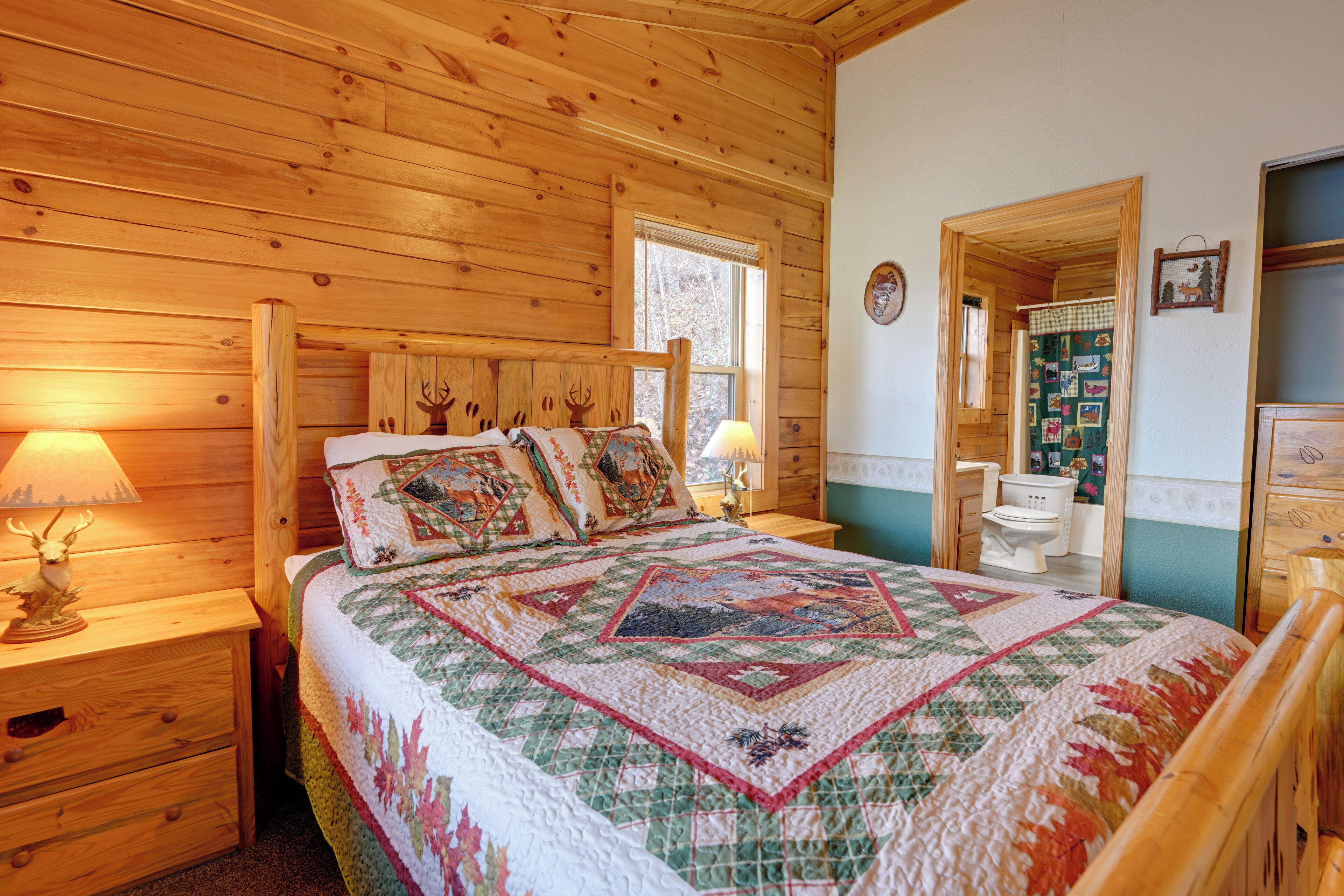 Une chambre à coucher située dans une cabane en bois, avec un lit, deux tables de chevet, une commode, une fenêtre et une salle de bain visible à travers une porte ouverte.