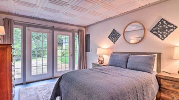 A bedroom with a large bed, a wooden nightstand, a mirror, and a window with a view of greenery.