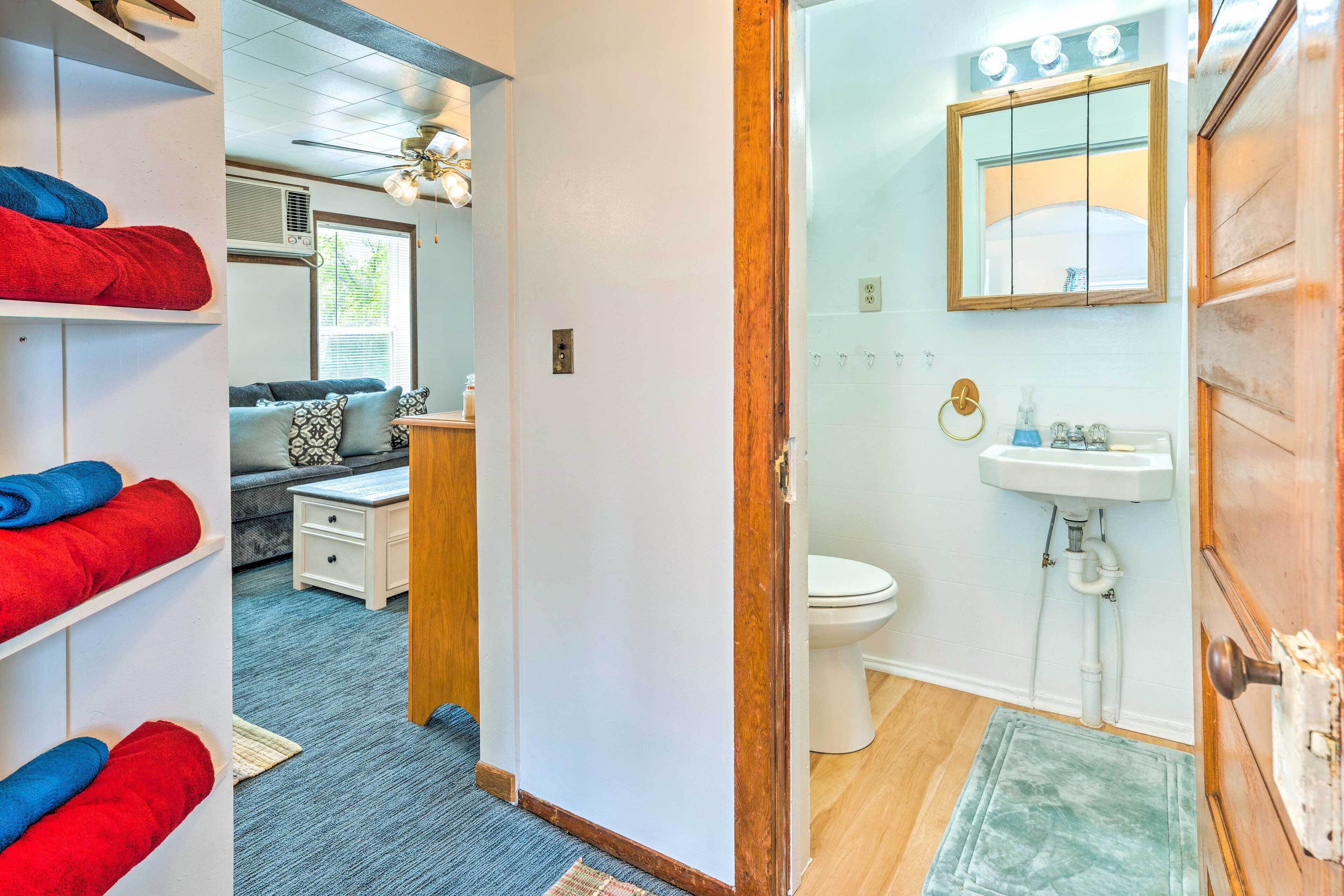 Un couloir mène à une salle de bain équipée de toilettes, d’un lavabo et d’un miroir. Une étagère blanche, sur laquelle sont posées des serviettes, se trouve dans la pièce adjacente, tout comme un ventilateur de plafond.