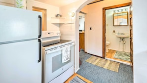 A compact kitchen with a refrigerator, stove, and microwave. There's a wooden door leading to a bathroom with a sink and mirror. - Historic Cabin Retreat: Steps to Okoboji Lake (Arnolds Park)