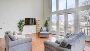 A spacious living room with grey sofas, a flat-screen TV, wooden flooring, and large windows offering a view of the outdoors.