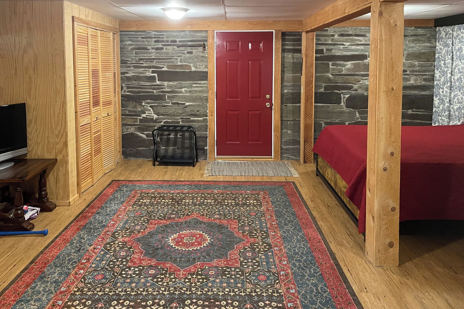 A room with a red door, a bed with a red cover, a wooden bench, a TV, and a patterned rug.