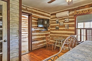 Quarto de cabana de toras com piso de madeira, uma cama, uma mesa de jantar com cadeiras, uma televisão fixada na parede e uma janela com persianas.