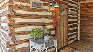 A rustic log cabin interior with a wooden door labeled "GRUENE," a small table with a potted plant, and a vintage-style light fixture. - 'gruene Cabin' - Pet Friendly New Braunfels Studio (New Braunfels)
