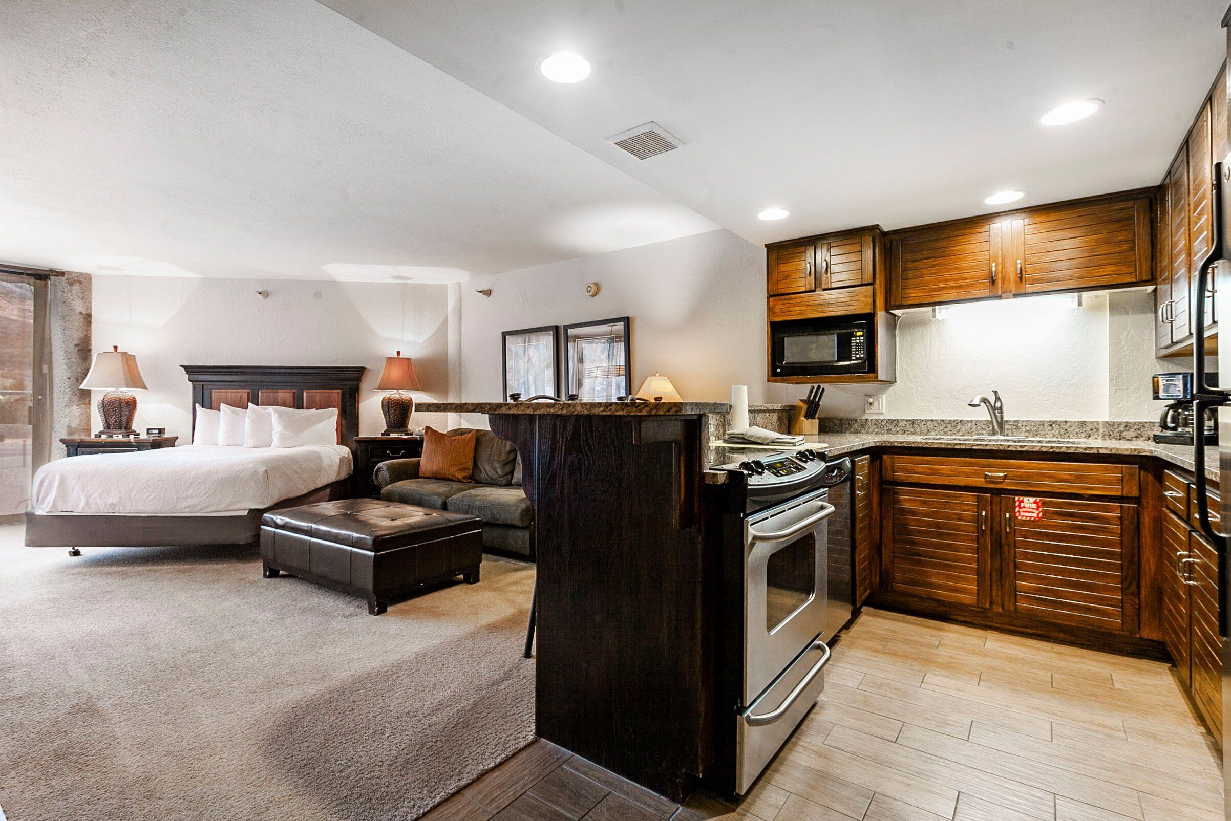 A modern hotel room with a large bed, a sofa, a dining area, and a kitchen with wooden cabinets and a microwave.