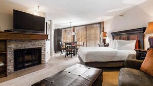 A hotel room with a large bed, a flat-screen TV mounted above a stone fireplace, a dining area with a table and chairs, and a cozy seating area with a cushioned bench and armchair.