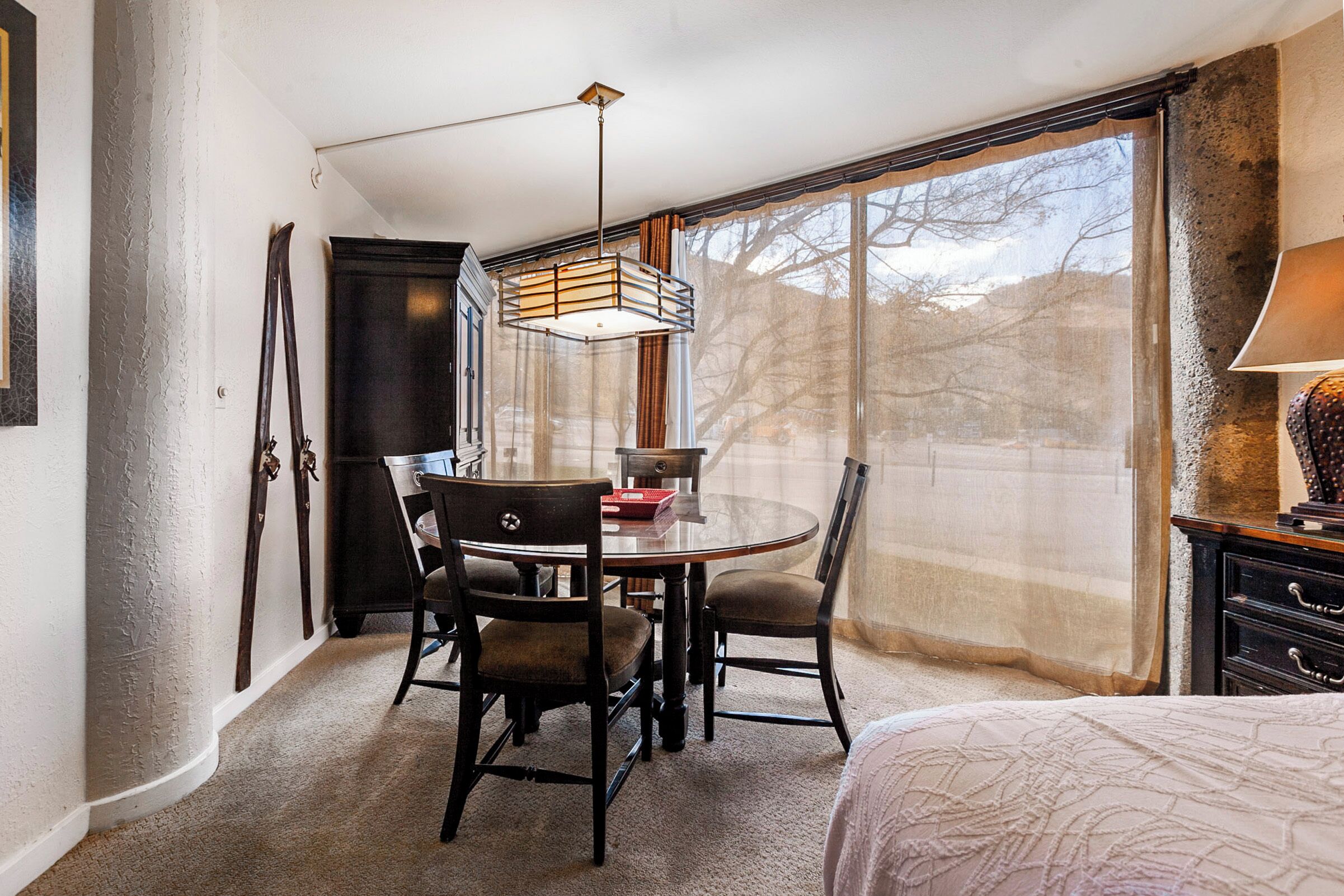 Une chambre d’hôtel avec une table de salle à manger ronde, deux chaises, un lit, une grande fenêtre avec des rideaux légers et une lampe sur une table de chevet.