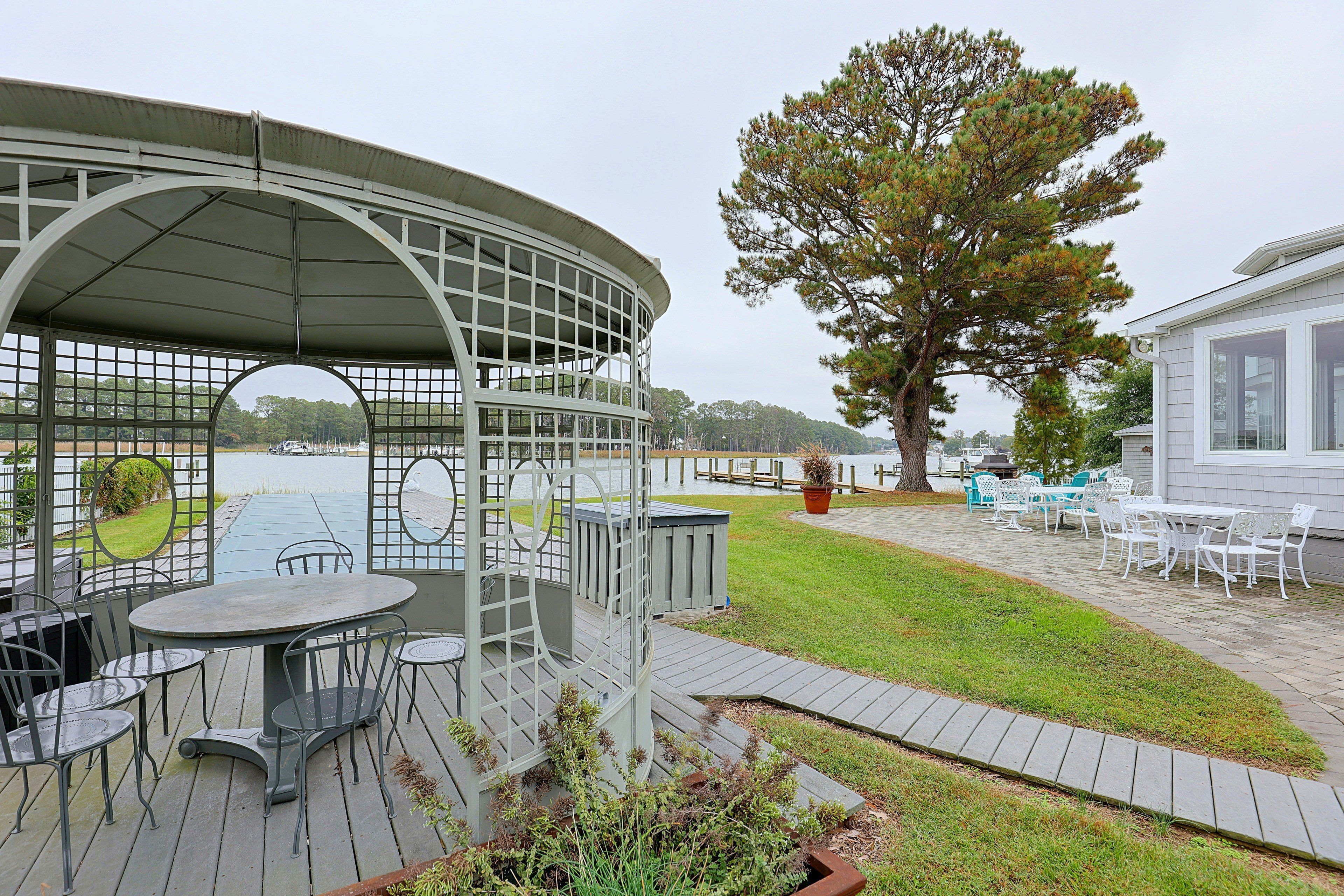 A covered outdoor seating area with tables and chairs, a large tree, and a view of a body of water.