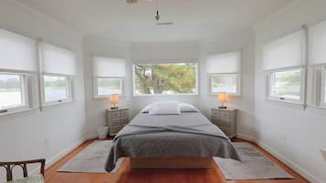 A bedroom with a large bed, two bedside tables with lamps, a view of trees through the windows, and a wooden floor.