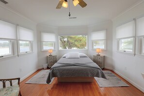 A bedroom with a large bed, two bedside tables with lamps, a view of trees through the windows, and a wooden floor. - Grasonville Home w/ Private Pool on the Water (Grasonville)