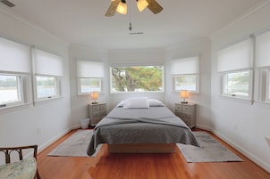 Un dormitorio con una cama grande, dos mesitas de noche con lámparas, vista a árboles a través de las ventanas y suelo de madera.