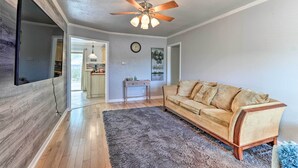A living room with a wooden floor, a beige sofa, a ceiling fan, and a flat-screen TV mounted on the wall. - 5 Mi to Fort Hood! Brooklyn House w/ Fenced Yard (Killeen)